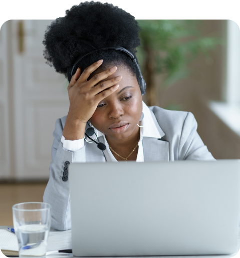 An employee engagement expert, a black woman in a grey blazer, looks stressed while working on a laptop, holding her forehead and wearing headphones.