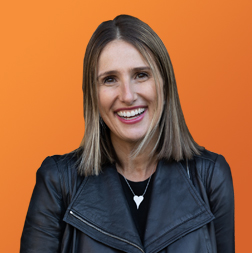 A woman in a black leather jacket smiling in front of an orange background.