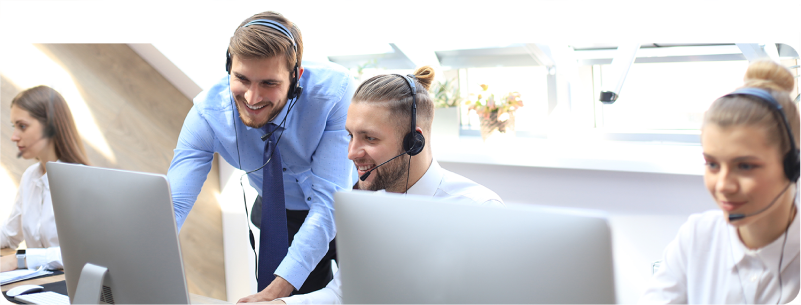 Two men with headsets, one standing and the other seated, collaboratively managing a multimodal customer experience, looking at a computer screen in a busy office setting.