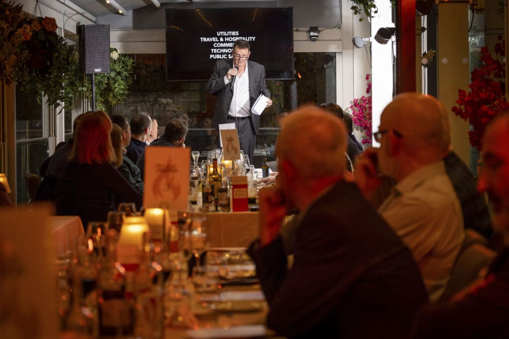 A man speaks into a microphone at a podium in front of an audience seated at dinner tables in a warmly-lit restaurant, with a screen displaying PR event topics behind him.