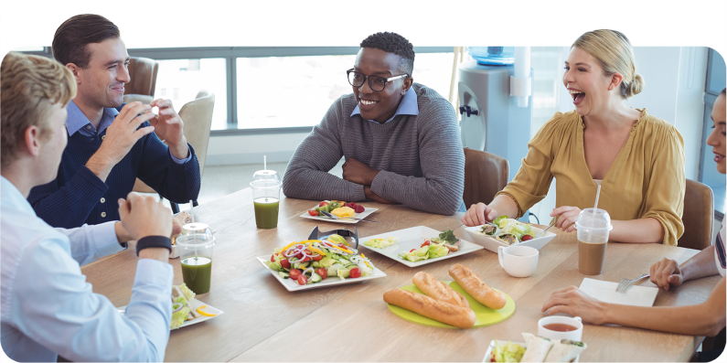 Group of customer service reps enjoying a lunch break around a table filled with healthy food, engaging in conversation and laughter in a bright office kitchen as a way to show appreciation.